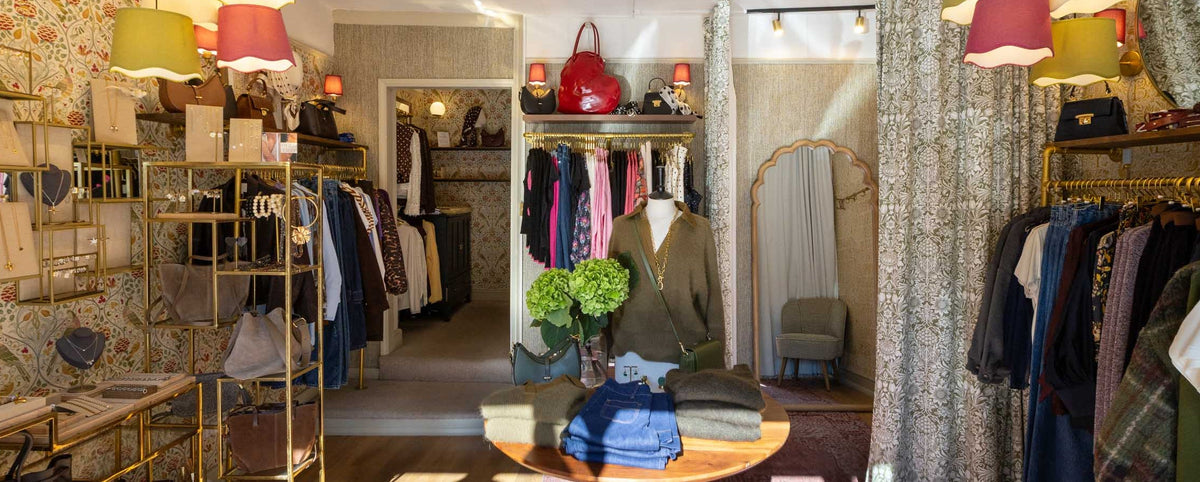 A boutique interior with clothing racks, patterned wallpaper, hanging pendant lights in pink, yellow, and cream, a central table with folded garments, and a mannequin wearing a green blazer.
